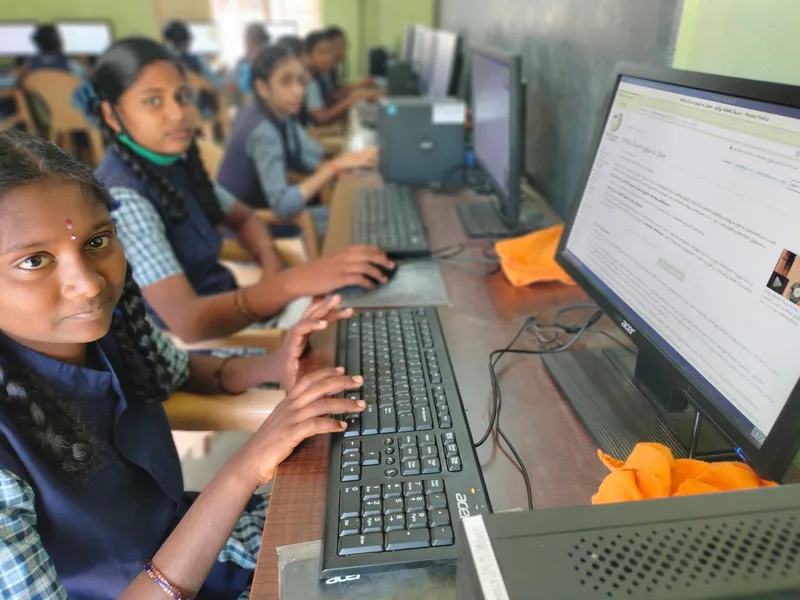 School students in uniform typing at desktop computers in a computer lab, one looking towards the camera