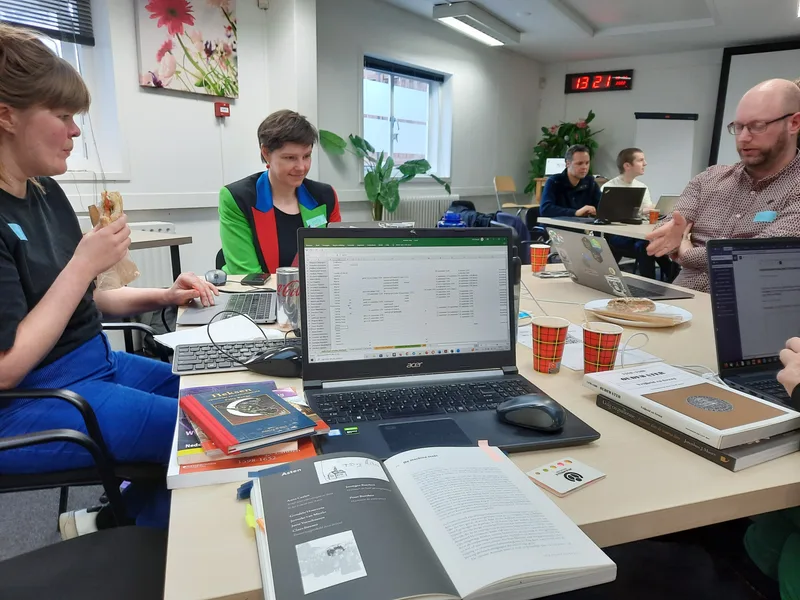 Hackathon participants around a table with laptops, books and coffee cups, discussing their work