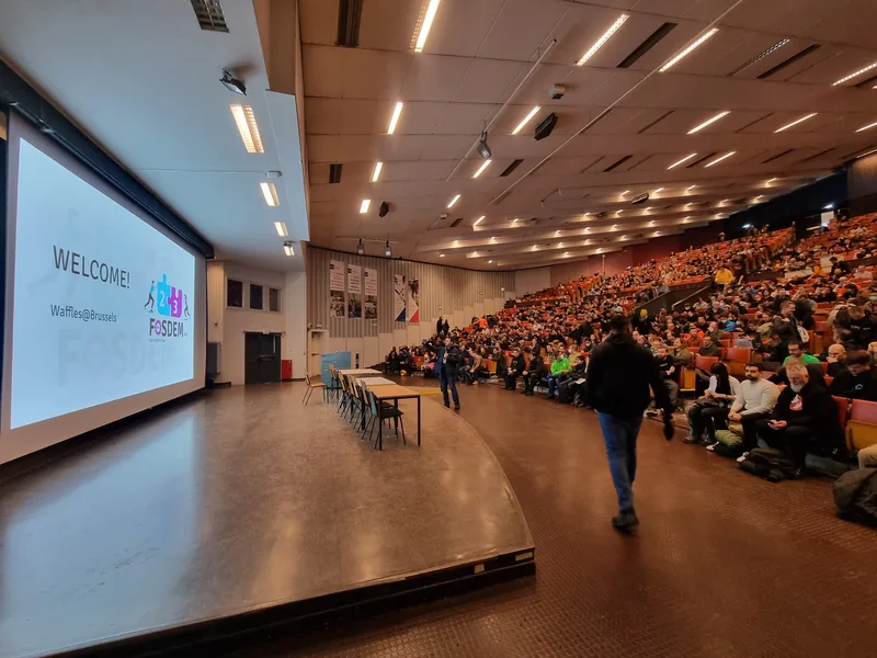 A large auditorium filling up before the Welcome to FOSDEM keynote, with the welcome slide on the projector screen