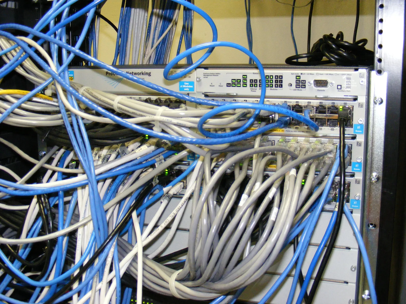 Gigabit network switch in a rack surrounded by a dense tangle of blue and grey Ethernet cables