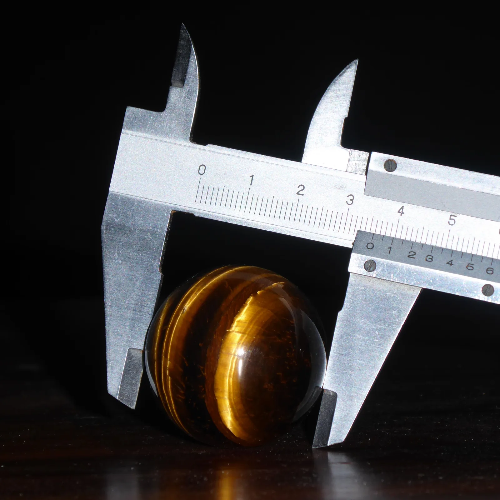Steel vernier caliper measuring the diameter of a polished tiger's eye sphere against a black background