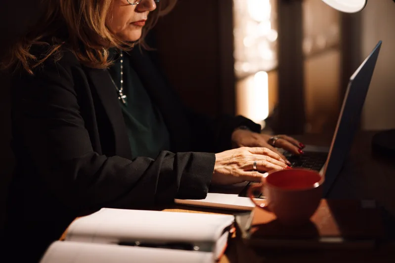 Woman typing on a laptop at a desk with an open notebook and a red coffee cup