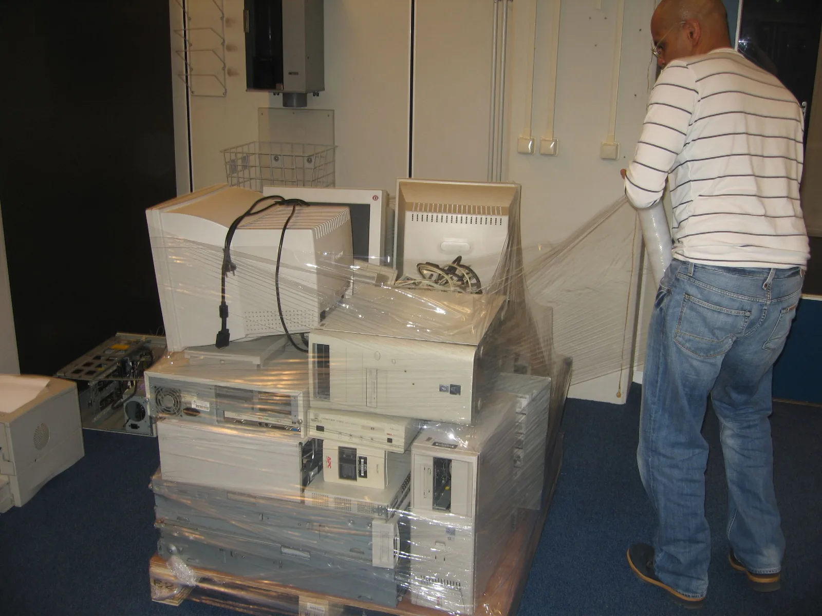 Old beige desktop computers and monitors stacked on a pallet and being wrapped in plastic film for recycling