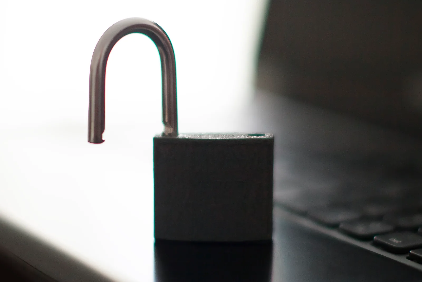 Open padlock standing on the edge of a dark laptop keyboard, backlit by a bright window