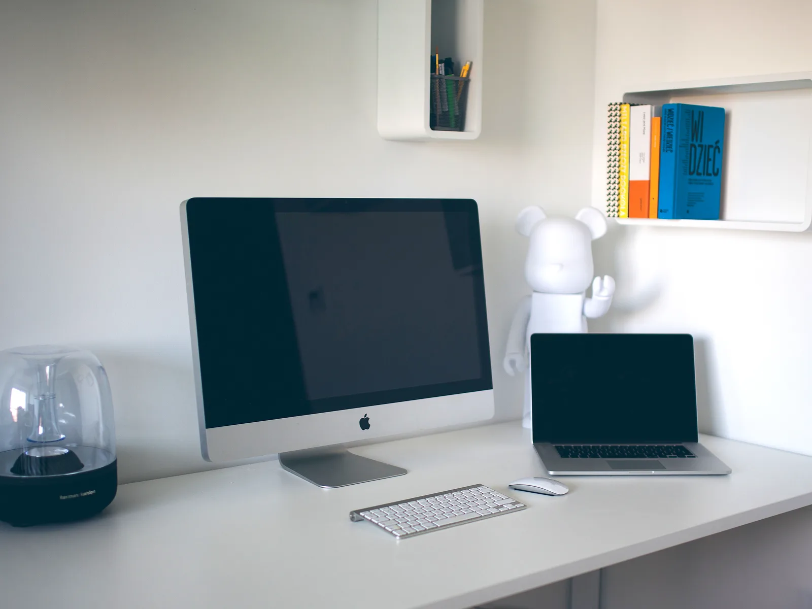 Tidy white desk with a desktop computer, a closed-screen laptop, keyboard and mouse, and books on a wall shelf