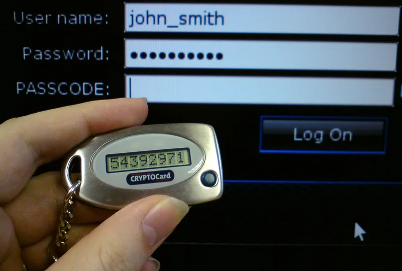 Hand holding a hardware token showing a one-time code in front of a login form with user name, password and passcode fields