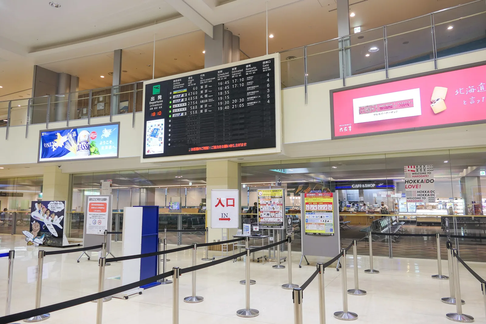 Airport terminal hall with rope barriers leading to a security check entrance marked IN, under a departures board