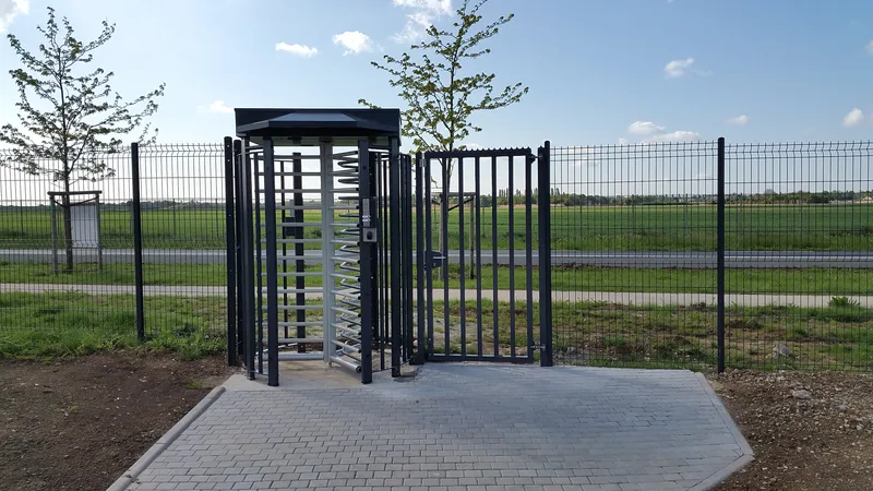 Full-height metal turnstile and gate set into a perimeter fence beside a grass field