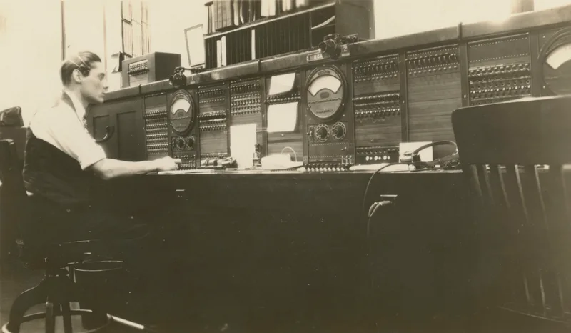 Young man seated at a large 1930 telephone switchboard with dials, meters and rows of jacks