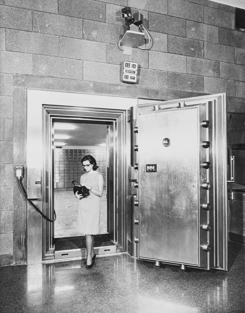 Black and white photo of a woman walking out through the open round-edged door of a bank vault