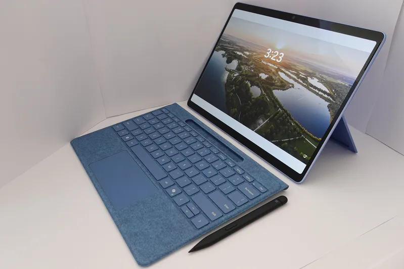 Light blue tablet-style laptop with a detachable fabric keyboard and a stylus on a white surface, its screen showing a lake landscape