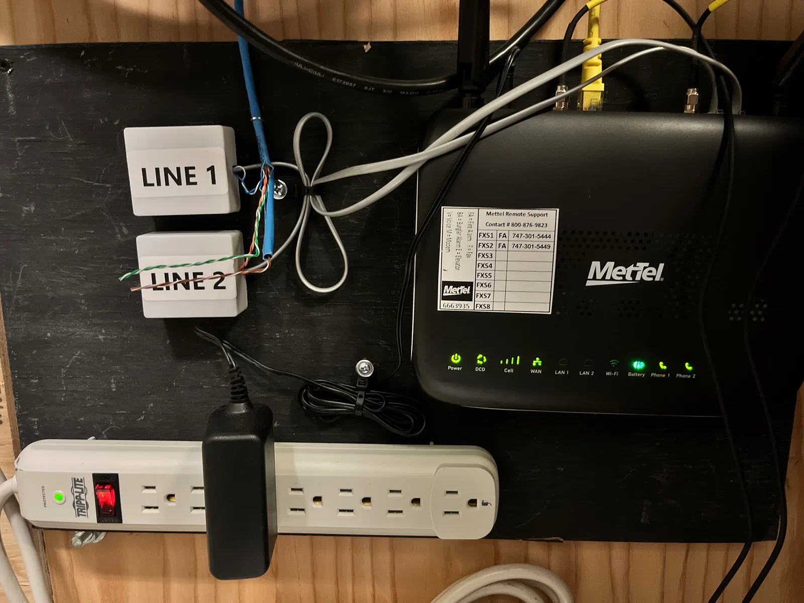 Wall-mounted network installation with a black router, two telephone line jacks, patch cables and a power strip
