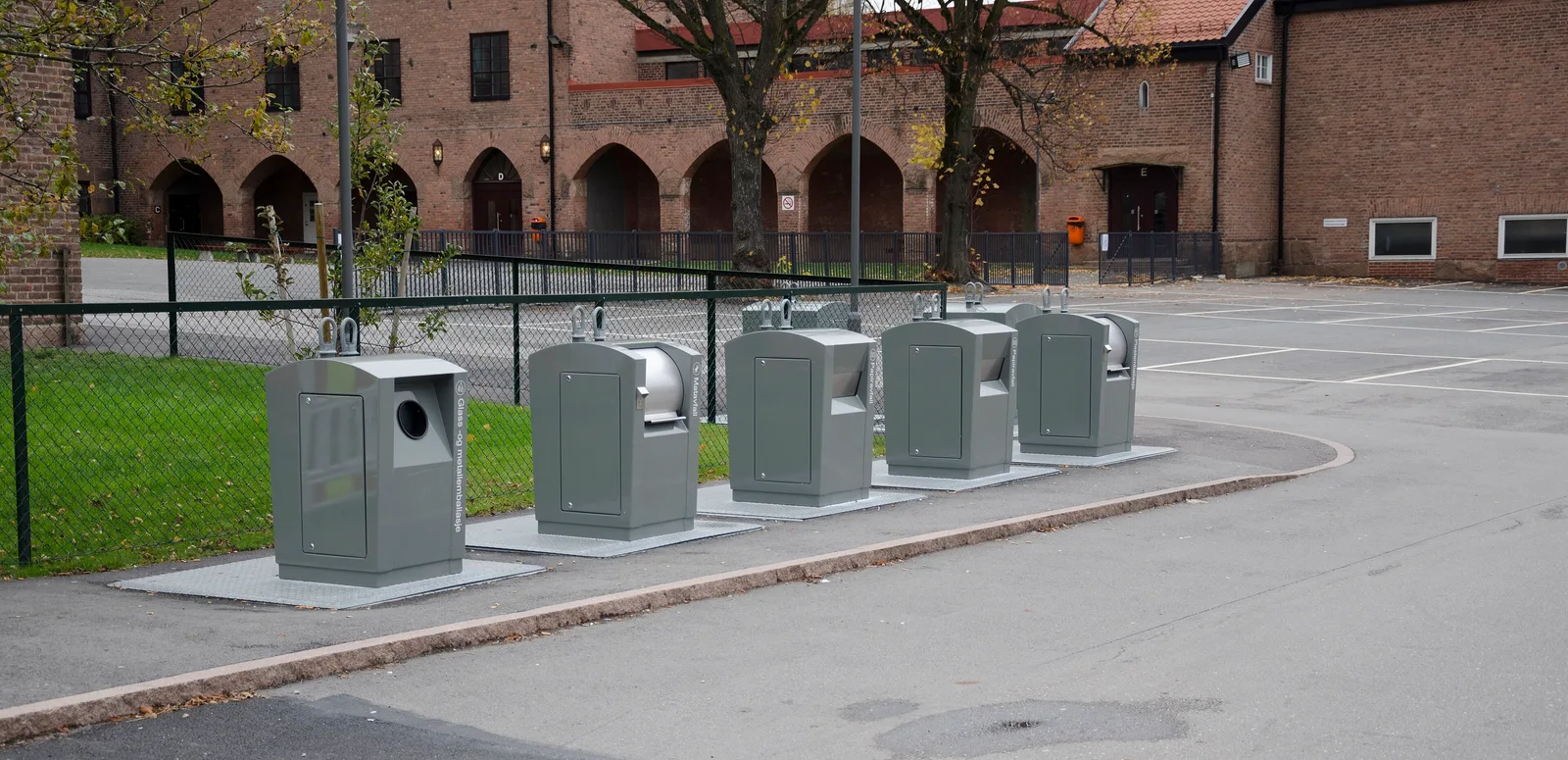 Row of grey underground recycling bins beside a pavement in front of a brick building in Drammen