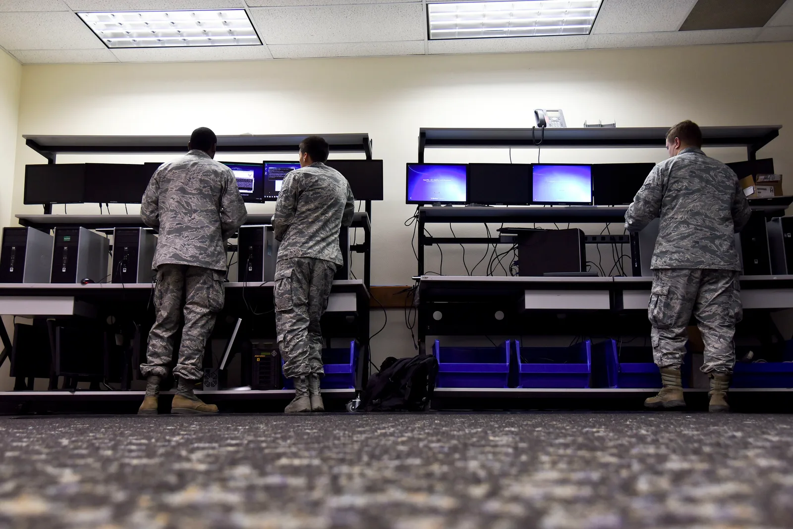 Three technicians in uniform working at rows of desktop computers on long workbenches