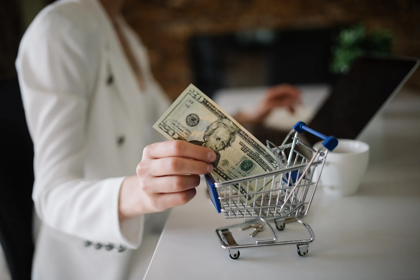 A woman places a banknote into a miniature shopping cart beside a laptop on an office desk
