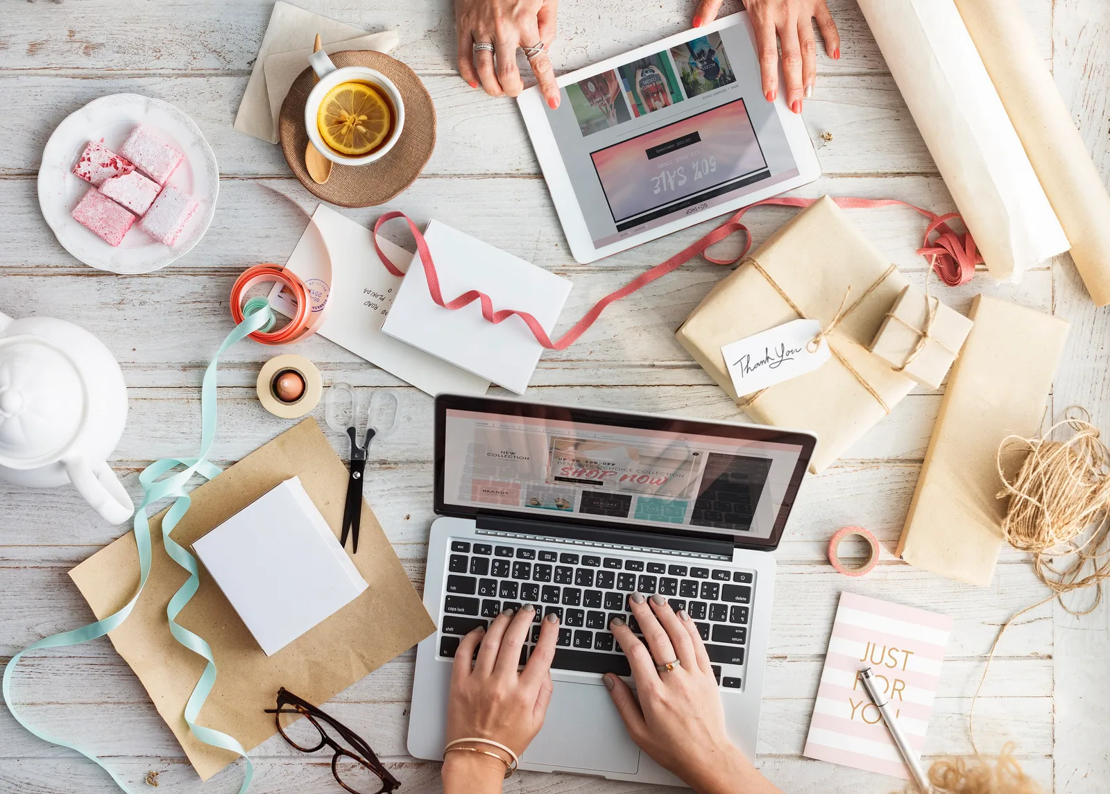 Overhead view of hands browsing a shop on a laptop and a tablet, surrounded by wrapped gifts and twine