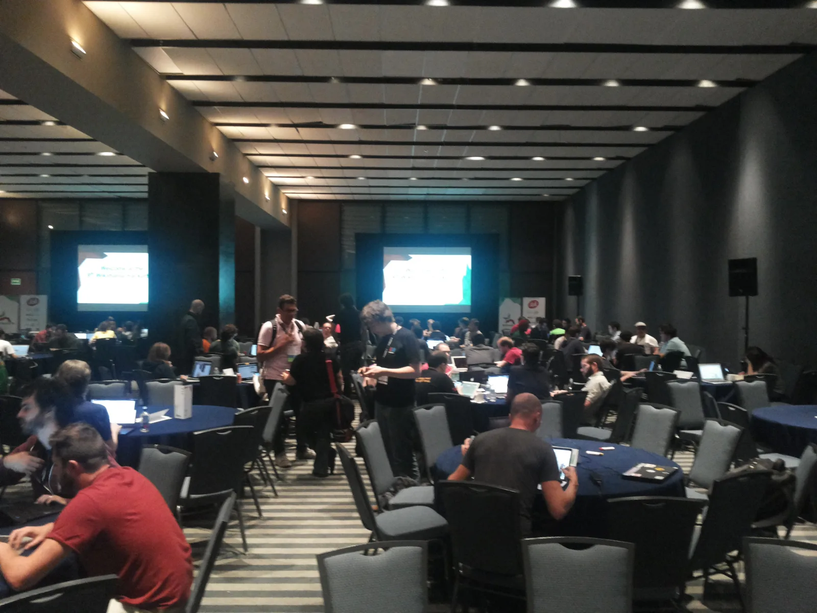 Large conference room with round tables where groups of developers work on laptops beneath projector screens