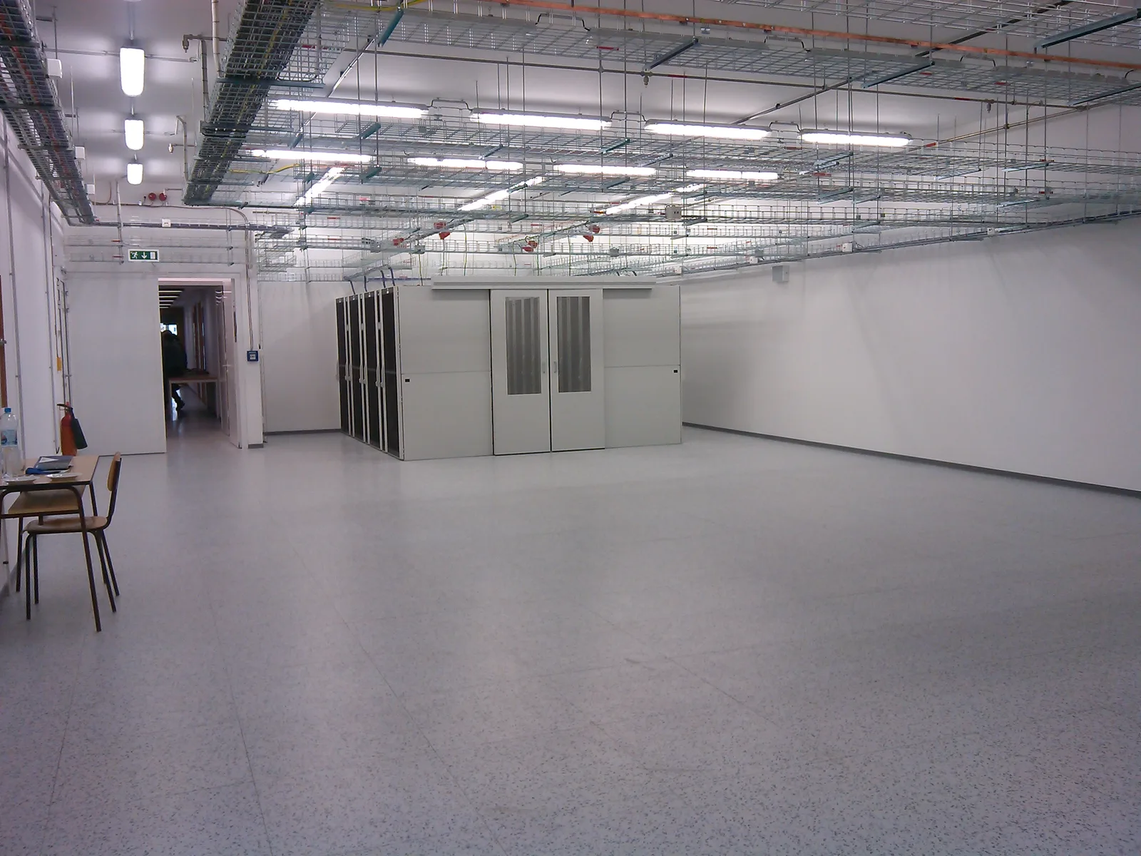 Large, mostly empty data centre hall with a single enclosed block of server cabinets under overhead cable trays