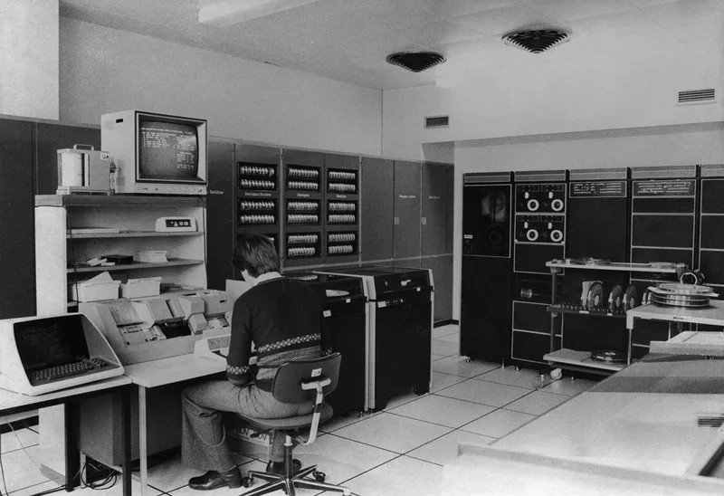Black-and-white photo of a historic computer room with an operator at a terminal beside tape drives and cabinets