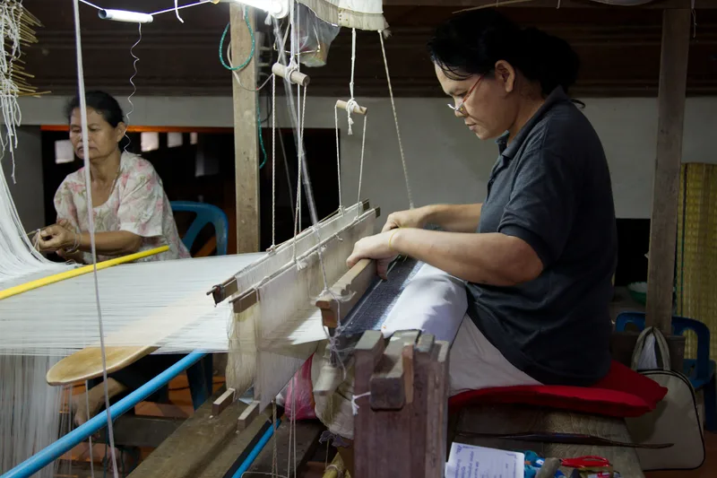 A woman weaving silk on a wooden hand loom while another woman prepares threads behind her in Ban Tha Sawang, Thailand