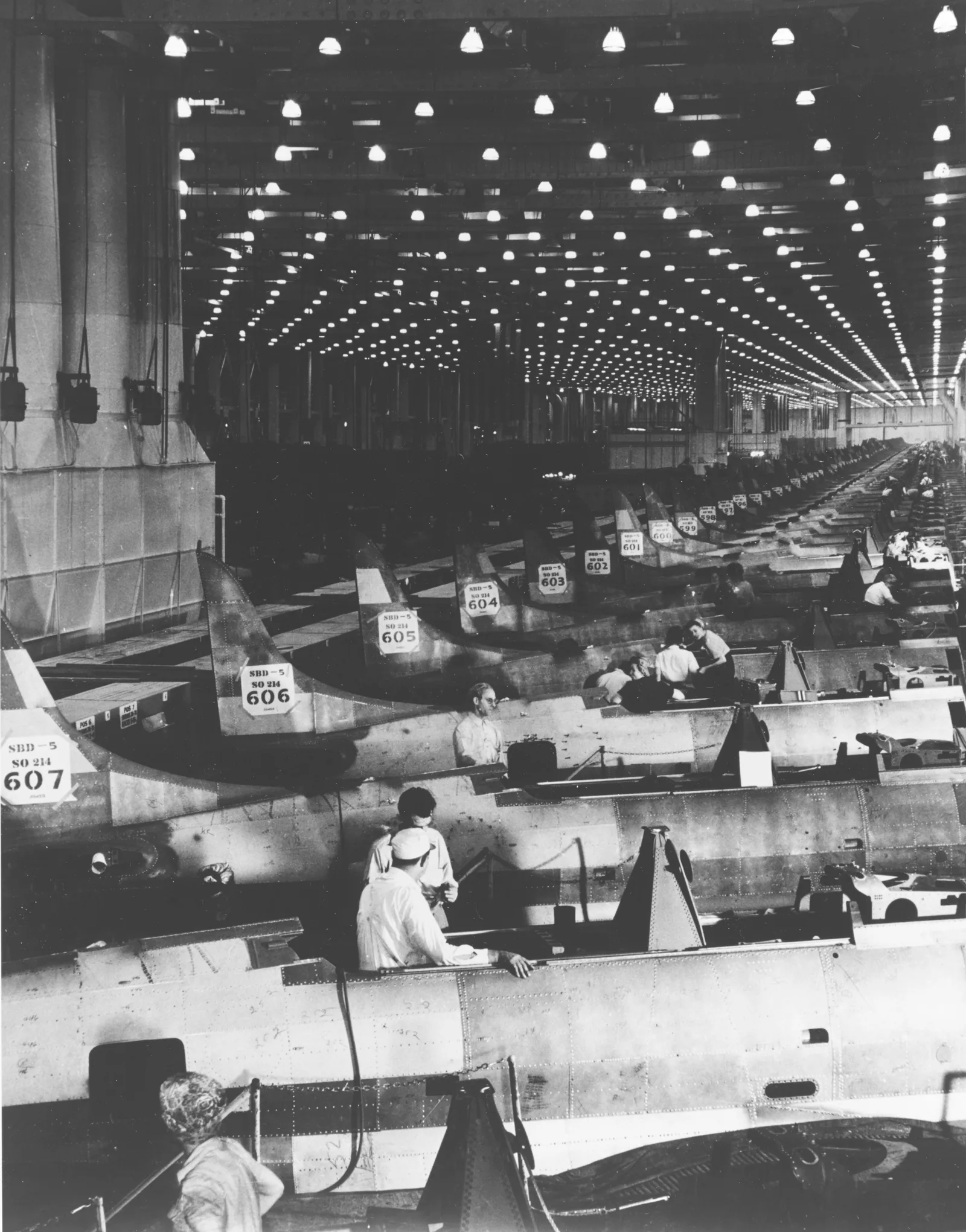 Black-and-white photo of a long aircraft assembly line in 1943, with workers on numbered fuselages under rows of ceiling lights