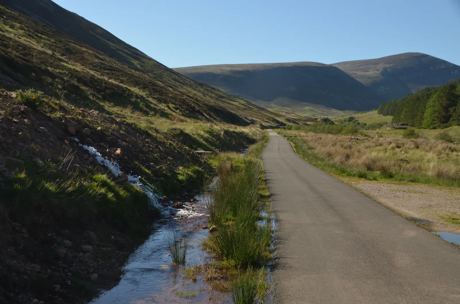 Water spilling from a hillside into a roadside ditch beside a narrow road through a green Highland glen under a clear sky