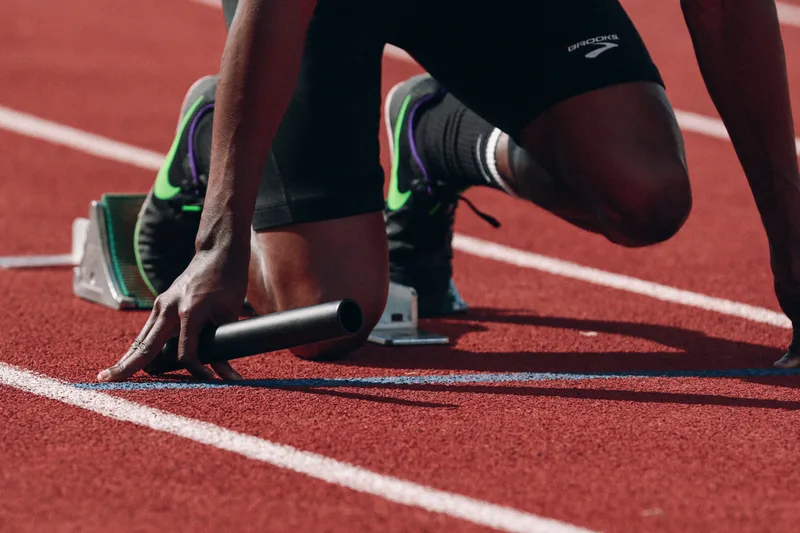 A sprinter crouched in starting blocks on a red running track, one hand resting beside a black relay baton