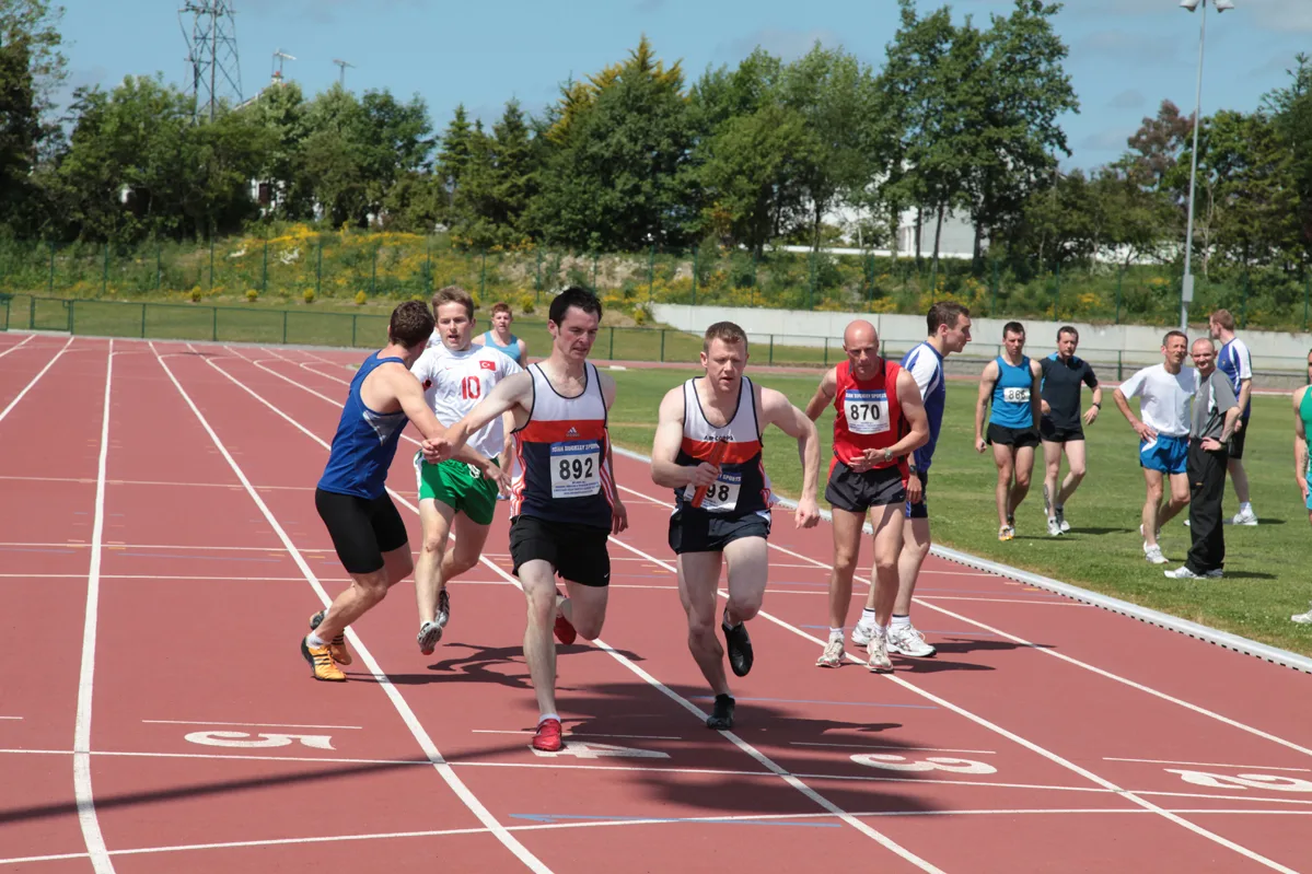Runners passing relay batons at a changeover zone on a red athletics track while other athletes wait on the grass