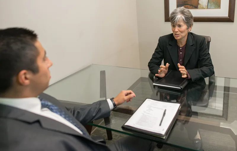 Interviewer gesturing while she speaks to a candidate across a glass table with a notepad and pen