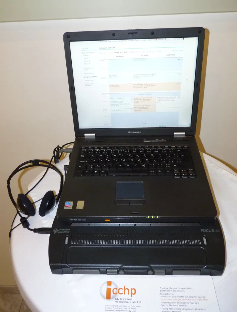 A laptop showing a conference programme web page, standing on a 40-cell braille display, with headphones plugged in beside it