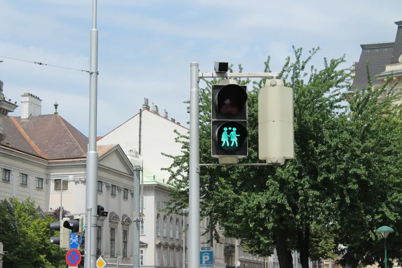 A pedestrian traffic light in Vienna showing a green signal with two figures, with trees and buildings behind it
