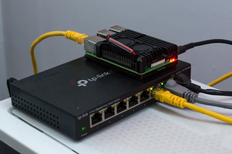 A Raspberry Pi 4 in a black heatsink case sitting on a small TP-Link managed network switch, with yellow and grey Ethernet cables plugged in
