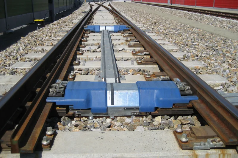 Close-up of a railway switch, with the movable rails, a blue drive housing between the sleepers and gravel ballast
