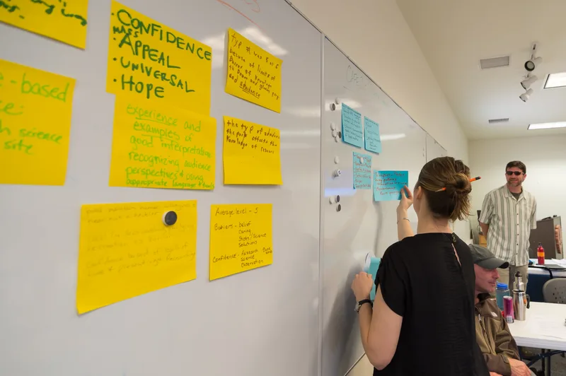 Woman adding a sticky note to a whiteboard covered in yellow and blue notes during a training workshop