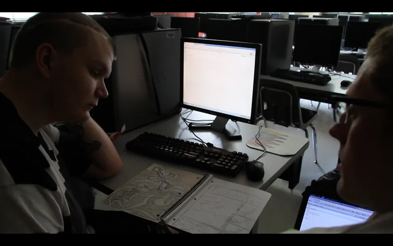 Two people at a computer desk discussing a paper sketch of a website layout