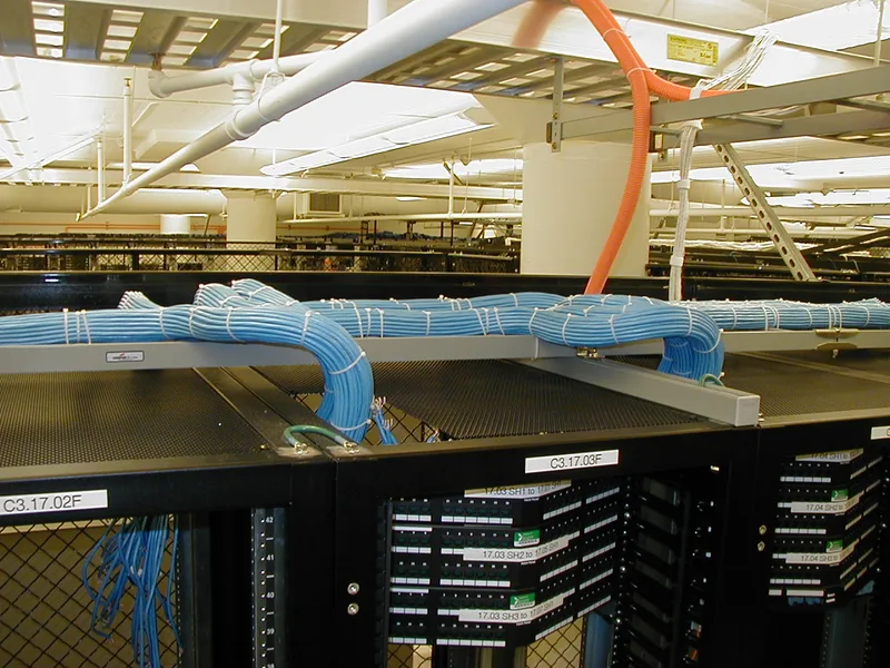 Bundles of blue network cables in an overhead tray running into labelled patch panels on black data centre racks