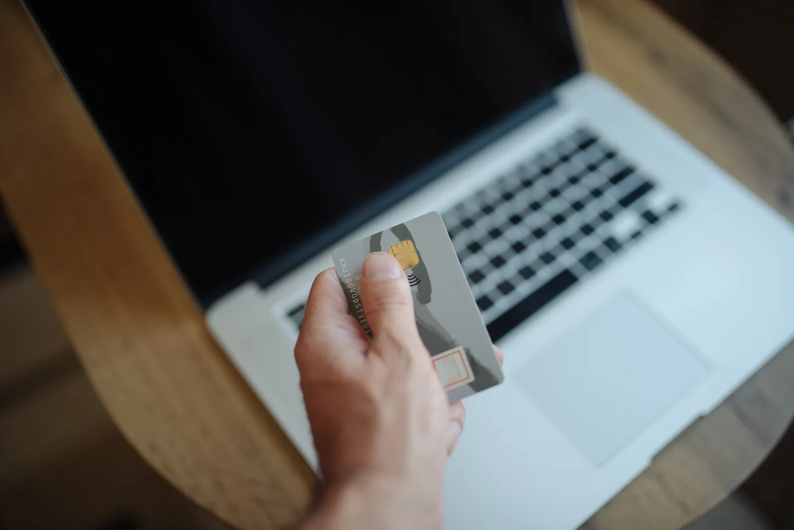 Hand holding a payment card in front of an open laptop on a wooden table