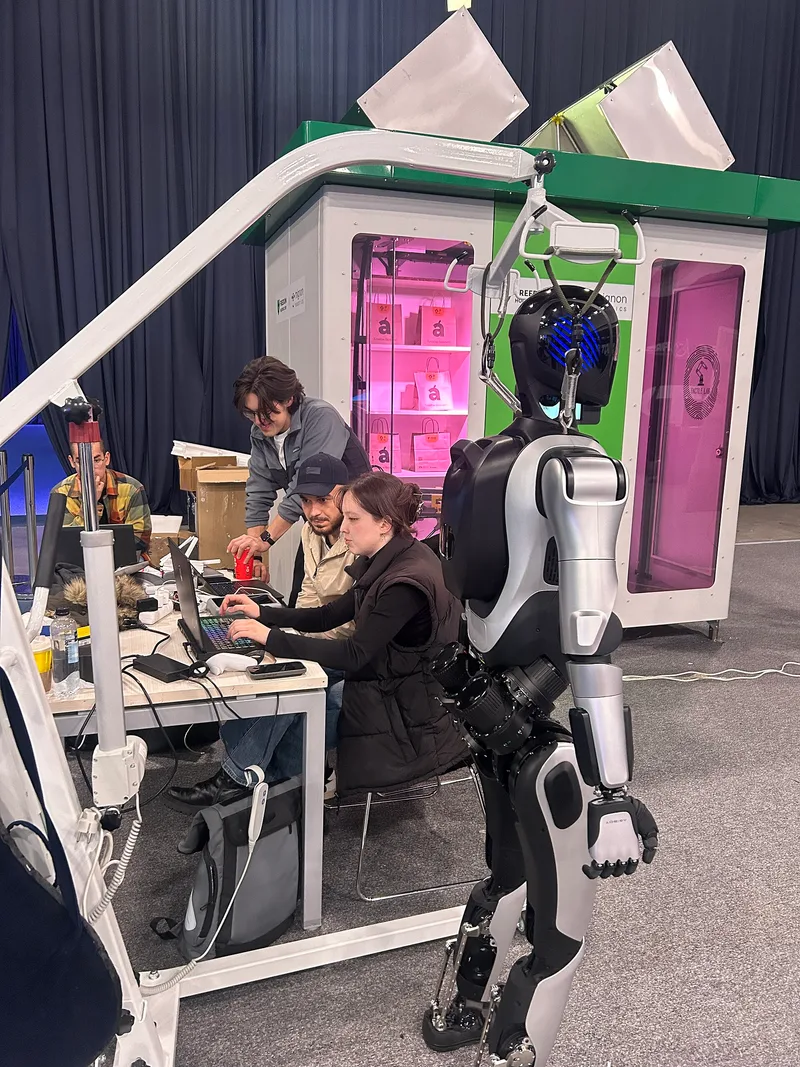 People working at laptops on a table next to a standing humanoid robot at an exhibition