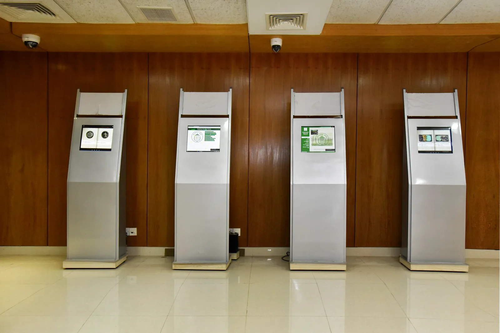 Four freestanding touchscreen information kiosks in a row against a wooden wall