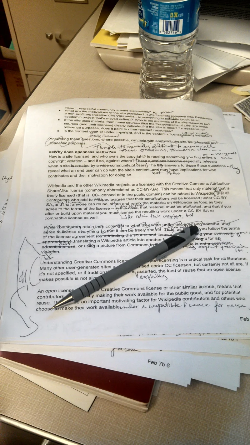 Printed manuscript pages on a desk covered in handwritten edits, strike-throughs and margin notes, with a pen lying across the top page