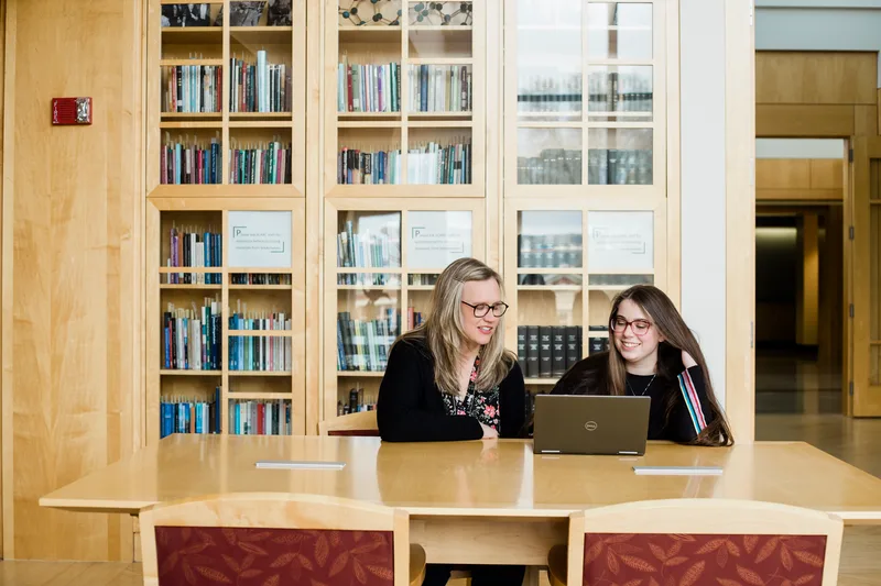 A librarian and a smiling student sit side by side at a library table looking at a laptop, with glass-fronted bookcases behind them