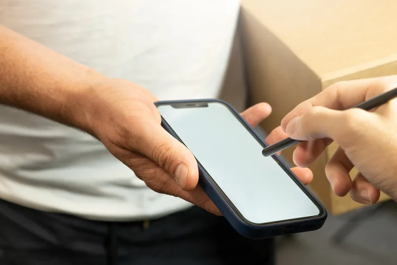 Close-up of a person signing with a stylus on a courier's smartphone while holding a cardboard parcel under one arm