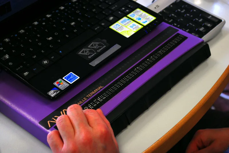 A hand resting on a purple 40-cell refreshable braille display with a row of raised pins, placed in front of a small laptop keyboard.