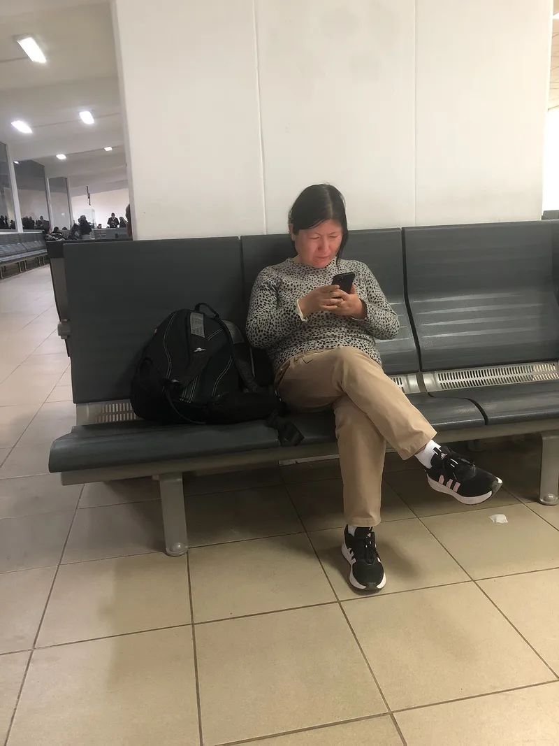 A woman sitting on a row of grey seats in an airport departure area, looking at her smartphone with a backpack beside her.